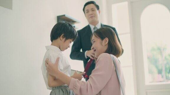 亚洲妈妈抱着她的小儿子准备送他去上学而亚洲爸爸在家里的客厅里背着儿子的书包准备去上学亲子关系或爱与纽带表达