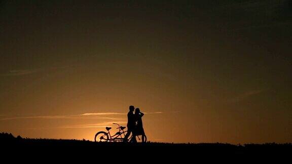 美丽的夫妇剪影与双人自行车行走在美丽的日落背景近距离
