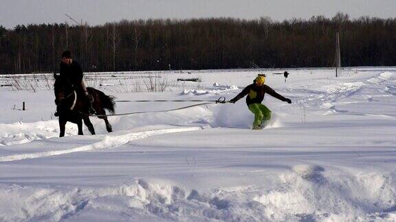 慢镜头:一个女孩骑着马疾驰一匹马用绳子拖着一个滑雪者滑雪者在雪堆中骑在滑雪板上女子骑师和男子滑雪板训练成对
