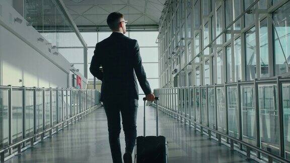一个穿着西装的年轻优雅的商人带着一个手提箱走在国际机场的慢镜头阳光从窗口射进来