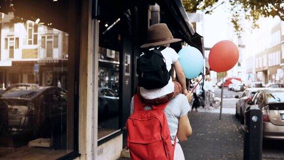母亲背着儿子沿街走女孩带着一个戴着帽子和两个气球的快乐小孩走着4k