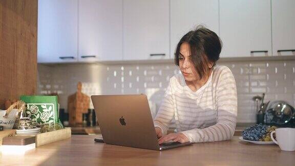 女性一边在厨房柜台上喝早餐咖啡一边用笔记本电脑远程工作