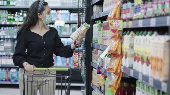 封锁后亚洲中年妇女挑选和购买果汁时在超市或商店购物和推着购物车时戴着医用口罩以防范冠状病毒疫情