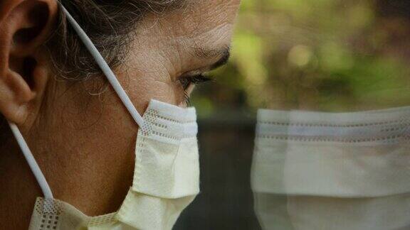 悲伤的成熟女人戴着外科口罩透过窗户看向别处