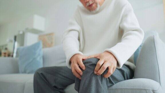 亚洲老年男子腿部受伤感到疼痛和痛苦有魅力的成熟男性患者坐在家里客厅的沙发上膝盖有问题医疗保健保险概念