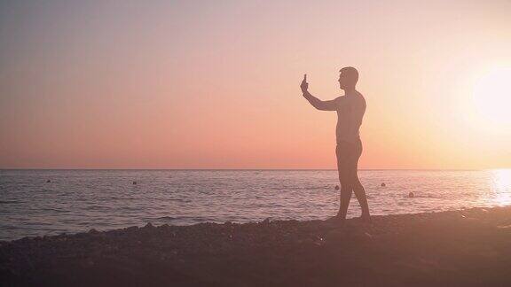 一个年轻人站在海岸上背景是海上的夕阳在日落的背景下男人在海滩上拍照