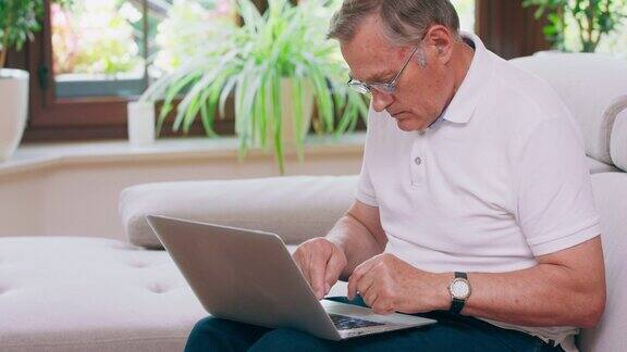 一位退休老人坐在家里的沙发上在笔记本电脑上打字