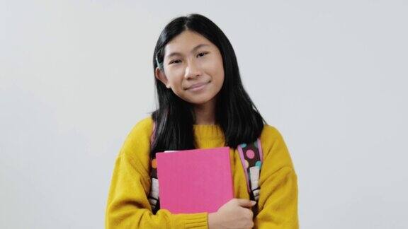 快乐的亚洲女学生穿着黄色毛衣拿着粉红色的书看着灰色背景的相机生活方式