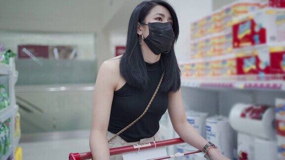 亚洲年轻女性黑色防护面罩推着购物车沿着杂货店通道妇女在锁定期间得到供应