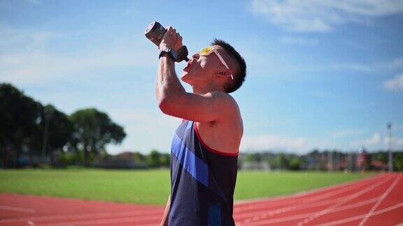 戴着太阳镜的男运动员早上在全天候的田径场上喝水