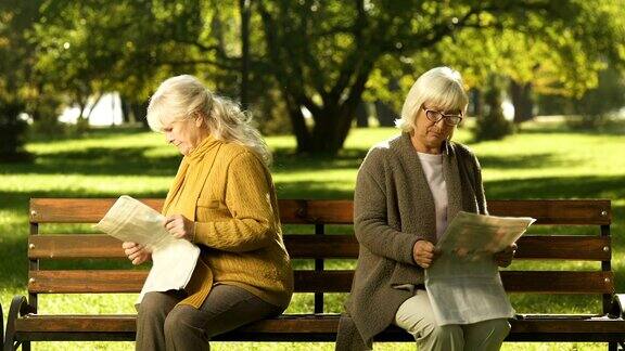 两位年长的女性朋友坐在公园的长椅上看报纸