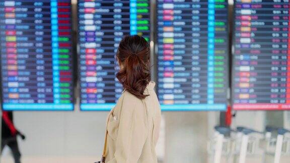 年轻的亚洲妇女拿着护照和登机牌在国际机场看航班信息板检查她的航班