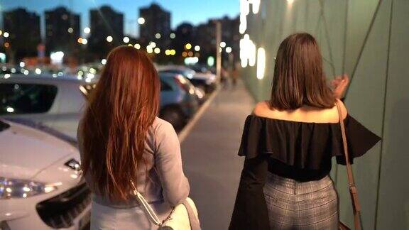 两名年轻女子在晚上走在人行道上的后视图