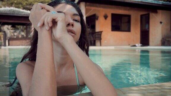 年轻迷人的比基尼女子站在别墅的泳池边看着镜头