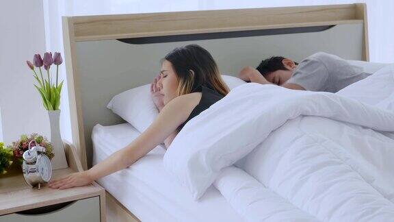 亚洲的一对年轻夫妇正躺在他们的卧室的床上闹钟在早上女人醒来看钟继续疲劳地睡