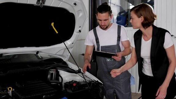 汽车服务男性汽车修理工站在打开发动机罩的汽车旁用平板电脑向客户解释问题