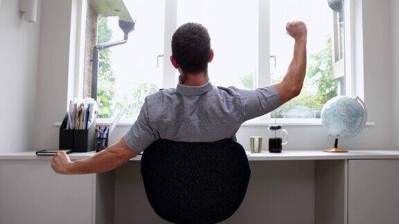 男人在家用电脑工作的后视图在家庭办公室伸展在书桌上