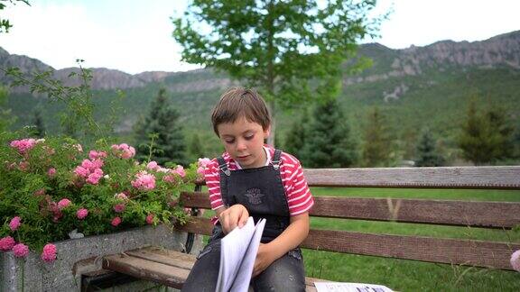 可爱的男孩坐在公园里的木凳上看书的肖像