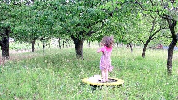 一个可爱的女孩在自然花园里跳舞小女孩在小蹦床上又跳又跳小女孩穿着花裙子戴着白色的帽子