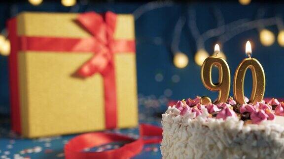 白色生日蛋糕90号金色蜡烛用打火机点燃蓝色背景用彩灯和黄色礼盒用红丝带绑起来特写镜头