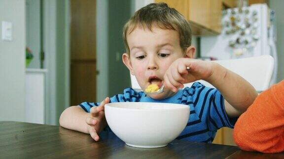 一个三岁的白人男孩在厨房的桌子上用勺子拿着碗吃通心粉和奶酪意大利面一边笑着一边做着傻乎乎的动作