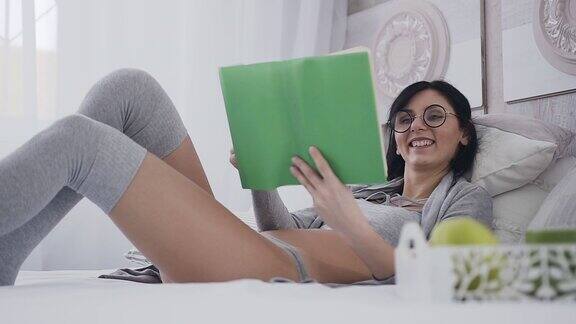 漂亮的小女孩戴着眼镜躺在卧室的床上她微笑着读着一本有趣的书穿着时髦的灰色衣服的女孩在早晨读一本有趣的书