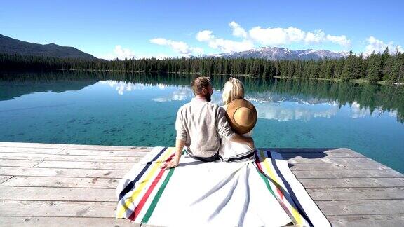 一对年轻夫妇坐在雄伟的山湖上的木栈桥上欣赏大自然