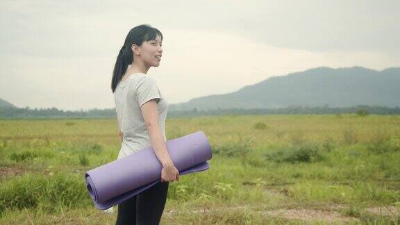 迷人的亚洲女人站着握着瑜伽垫在美丽的山景在早上