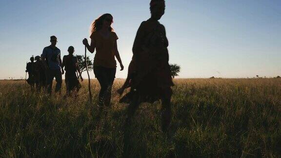 博茨瓦纳一群游客在Makgadikgadi草原上与身穿传统服装的桑族人布希曼人一起在丛林中漫步身后是夕阳