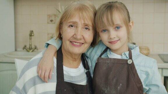 在厨房里穿着围裙的祖母和孙女微笑着看着照相机