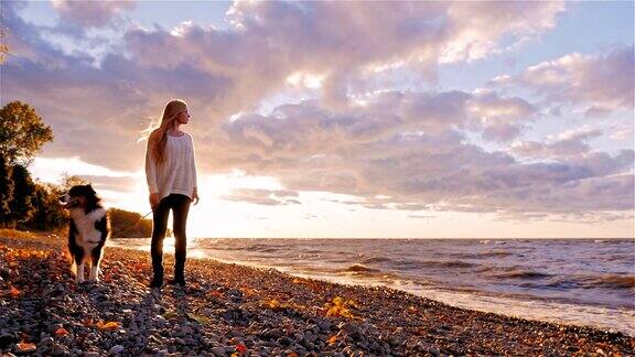 一名年轻女子带着一只狗站在湖边或海边夕阳西下遥望远方做梦