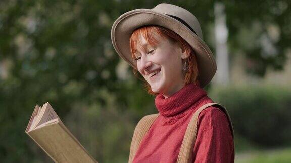 年轻迷人的红头发女人戴着帽子和毛衣在自然公园看书秋天凉爽的一天