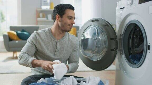穿着灰色牛仔裤和衬衫的英俊微笑的年轻人坐在家里的洗衣机前他把脏衣服装在洗衣机里明亮宽敞的客厅现代的室内设计